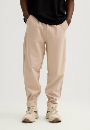 Persona con pantalones beige de corte ancho con las manos en los bolsillos y zapatillas beige, de pie frente a un fondo liso.