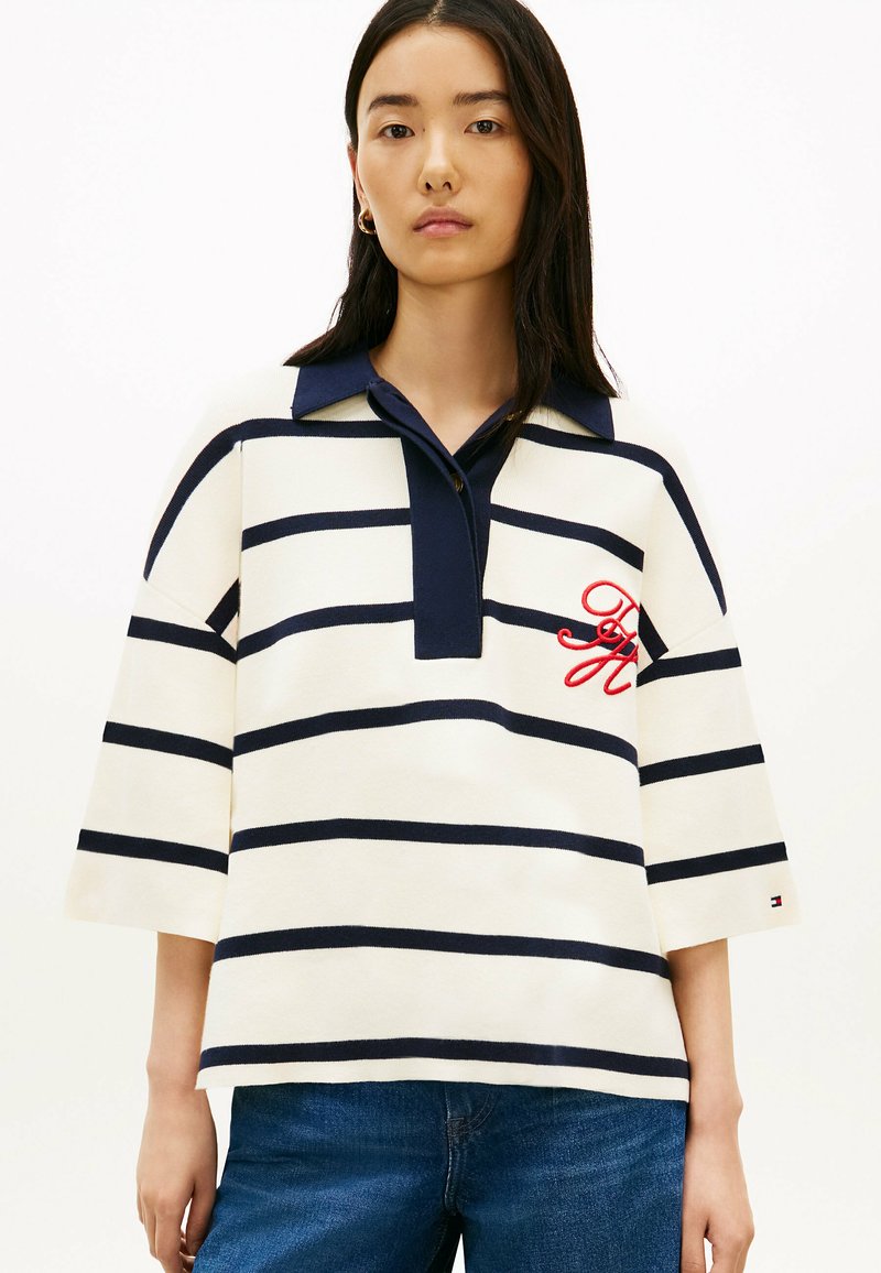 Mujer joven con cabello largo y oscuro que lleva una camisa de rayas blancas y azul marino con mangas a medio brazo y cuello azul, combinada con jeans azules.