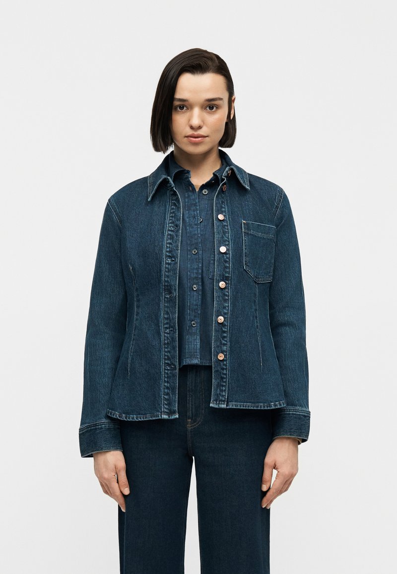 Νεαρή γυναίκα που στέκεται με το βλέμμα μπροστά, φορώντας σκούρο τζιν μπουφάν πάνω από ένα κουμπωτό τζιν πουκάμισο και τζιν παντελόνι που ταιριάζει.