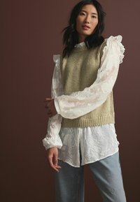 Strik en olivenfarvet vest over en hvid tekstureret bluse med volanger på ærmerne, parret med lyseblå jeans med udsmykning. Mørk baggrund.