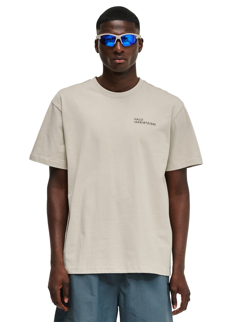 Jonge man met blauwe reflecterende sportzonnebril, beige oversized t-shirt met de tekst "HALO" en blauwe short, staand tegen een witte achtergrond.