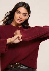 Burgundowa dzianinowa sweter z prążkowanym kołnierzem i mankietami, cechujący się luźnym krojem i teksturowanym materiałem. Modelka poprawia rękaw.