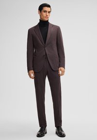 Dunkelvioletter Anzug mit zwei-Knopf-Blazer, mit Revers und flachen Vorderhosen. Über einem schwarzen Rollkragenpullover getragen; kombiniert mit schwarzen eleganten Schuhen.