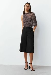 Brown polka dot sheer top with draped design, paired with high-waisted black culottes and black strappy heels. Gold bracelet accent.