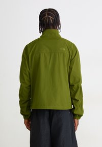 Man with braided hair wearing a green The North Face jacket and black pants, standing facing away against a plain background.