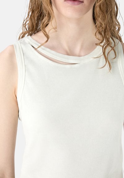 Femme aux cheveux bouclés portant un débardeur sans manches écru côtelé avec un petit détail en découpe près de l'encolure.