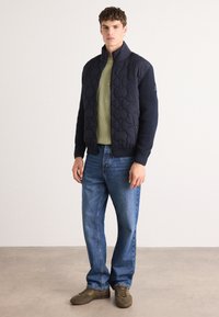 Marine quilted jakke med ribbet ermer, grønn strikkegenser, blå jeans og brune sko. Teksturert stoff og kontrastmaterialer.