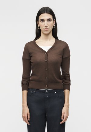 Žena s dlhými tmavými vlasmi, ktorá nosí zlaté kruhové náušnice, hnedý zapínací kardigán cez biely top a tmavomodré džínsy, stojaca pred jednofarebným pozadím.