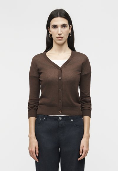 Woman with long dark hair wearing gold hoop earrings, a brown buttoned cardigan over a white top, and dark blue jeans, standing against a plain background.