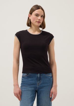 Vrouw draagt een zwarte mouwloze top met witte stiknaden en een blauwe spijkerbroek, staand tegen een effen lichte achtergrond.