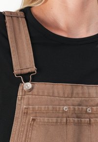 Bruine denim tuinbroeken met verstelbare banden, metalen clips en knopenaccenten. De stof heeft een gestructureerde afwerking en voorzakken. Zwarte shirt zichtbaar.