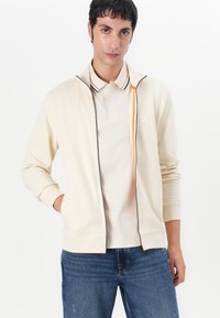 Felpa cremosa con zip alta, cerniera grigia e accenti arancioni. Indossata sopra una polo beige, abbinata a jeans blu.