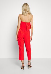 Combinaison rouge à design bustier, jambes fuselées et détail de nœud à l'arrière. Comprend une fermeture éclair et une texture de tissu lisse.