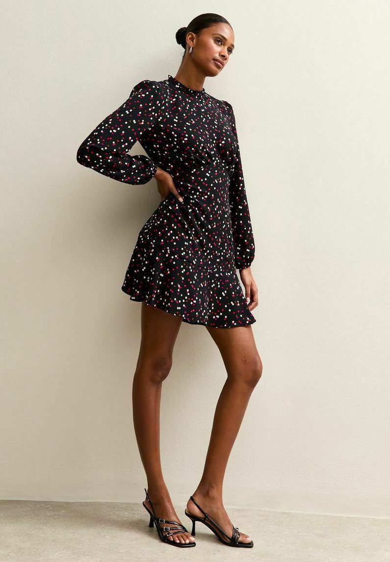 Schwarzes Kleid mit langen Ärmeln, das mit einem verstreuten Punktemuster in Rosa und Weiß verziert ist, und einem ausgestellt Rock. Getragen mit schwarzen Absatzsandalen.