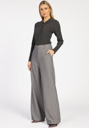 Vrouw met lang blond haar, gekleed in een donkergrijze geribbelde trui en hoog-waist grijze wijde broek met bruine hoge hakken.