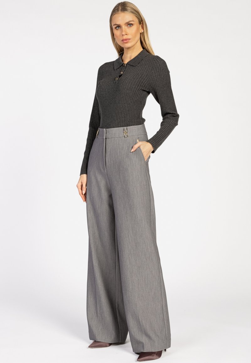 Vrouw met lang blond haar, gekleed in een donkergrijze geribbelde trui en hoog-waist grijze wijde broek met bruine hoge hakken.