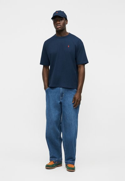 Άνδρας με μπλε καπέλο και μπλούζα με μικρό κόκκινο λογότυπο, χαλαρά μπλε τζιν και παπούτσια πράσινα και πορτοκαλί, στέκεται μπροστά σε απλό λευκό φόντο.