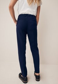 Pantaloni su misura color blu navy in tessuto liscio, fit slim e tasche laterali sottili, abbinati a scarpe nere slip-on.