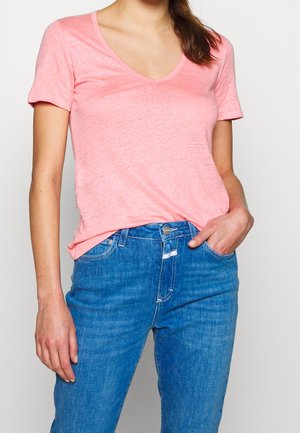 Vrouw draagt een lichtroze V-hals T-shirt met korte mouwen, lichtjes ingestopt in een middelblauwe spijkerbroek, met één hand in de zak.