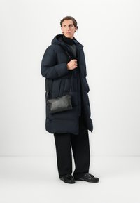 Mand iført lang sort pufferjakke, sort turtleneck, brede sorte bukser, sorte loafers og bærende en sort crossover-taske, stående mod en hvid baggrund.