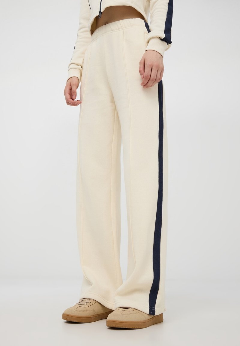 Pantaloni della tuta color crema realizzati in materiale morbido, con una striscia laterale blu navy, design a gamba larga e vita elastica.