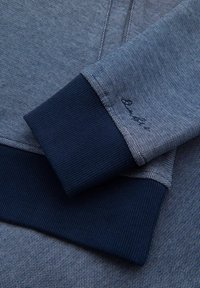 Tejido azul texturizado con un patrón sutil, con puños acanalados en azul oscuro. Se puede ver el detalle de la costura en la manga.