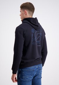 Marineblaues Kapuzenshirt mit Kordelzug und gerippten Bündchen, mit einem geometrischen Design in Blau auf dem Rücken. Kombiniert mit blauen Jeans.