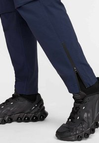 Marineblauwe sportbroek met een ritsdetail bij de enkels, gecombineerd met zwarte sneakers met een gestructureerde bovenkant en een ronde zool met vijf bubbels.