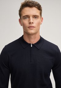Marineblaues Polohemd mit langen Ärmeln, Reißverschlusskragen, klassischem Kragen und glatter Textur. Der Stoff scheint leicht und figurbetont zu sein.