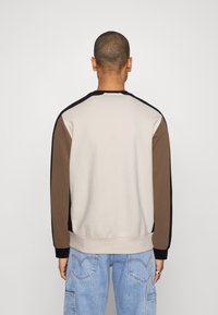Man draagt een color-block sweatshirt met een beige lijf, bruine mouwen, zwarte accenten en lichtblauwe jeans, kijkt naar achteren tegen een effen achtergrond.