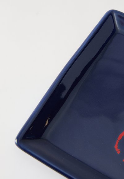 Négyzet alakú, fényes tengerészkék kerámia tányér hajlított szélekkel, egyik sarkában piros absztrakt mintával. Simább textúrájú, fényvisszaverő felülettel.