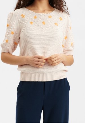 Mujer con suéter de punto corto de manga corta color rosa claro con detalles de flores naranjas y pantalones azul oscuro, manos entrelazadas en la cintura.