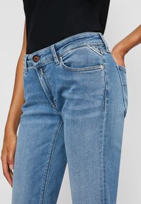 Kvinne iført lyseblå høytlivsjeans med en knapp foran, synlige sømmer og en liten trekantet merkeklipp på høyre lomme.
