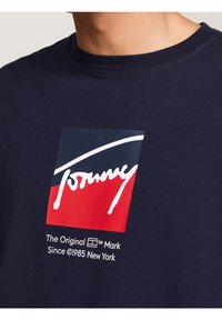 Tmavě modré bavlněné tričko s obdélníkovým logem v červené, bílé a námořnické modři. Text obsahuje "Tommy" a "The Original Mark Since 1985 New York."