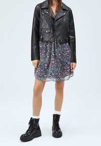 Svart läderjacka i bikerstil med kortärmar över en blommig, flerfärgad klänning med kort ärm. Svarta snörkängor och vita strumpor bärs.