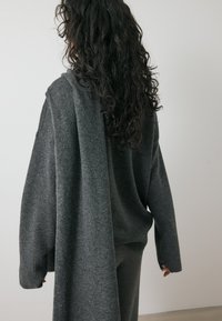 Temno siva oversize pulover z mehkimi teksturami, z dolgim drapiranim hrbtom, sproščenim krojem in rebrastimi manšetami, nošen z usklajenimi hlačami.