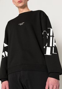 Černá mikina s malým bílým logem "Calvin Klein Jeans" na hrudi a výrazným bílým textem na rukávech. Svůdné manžety a lem.