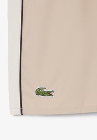 Panneau de tissu beige et blanc avec des détails en bordure noire et un logo crocodile vert brodé près de l'ourlet.