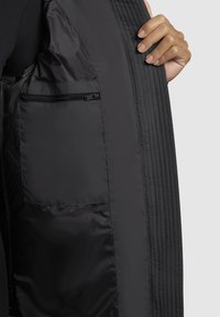Una mano tiene aperto un cappotto nero, rivelando una tasca interna con cerniera su un rivestimento di tessuto liscio, contro uno sfondo grigio.