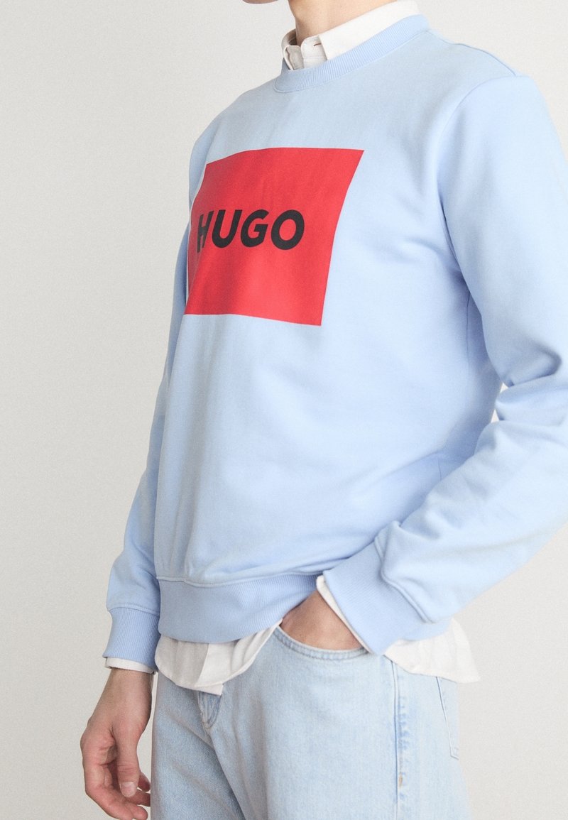 Sweat-shirt bleu clair en coton, avec un grand graphique rectangulaire rouge portant le texte « HUGO » en lettres noires. Poignets et ourlet côtelés.