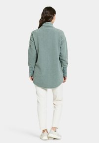 Fuzzy ljus teal-pullover med hög krage, långa ärmar och rundad nederkant. I kombination med vita byxor och ljusa sneakers.