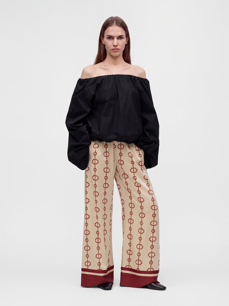 Femme portant un chemisier noir épaules dénudées à manches longues et un pantalon large beige avec un motif chaîne géométrique rouge, debout sur fond blanc.
