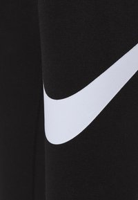 Tissu noir avec un grand logo en forme de swoosh blanc de Nike. La texture est lisse, typique du coton ou d'un mélange de coton.