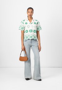 Kortebloemige witte overhemdd met groene bloemenborduursels en een gegolfde zoom, gecombineerd met lichtblauwe flared jeans en een kleine bruine handtas.