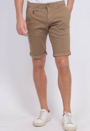 Homme portant un short marron retroussé avec fermeture bouton et zip, debout avec des baskets blanches sur fond uni.
