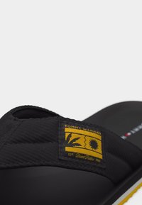 Μαύρο σανδάλι slide Tommy Hilfiger με κίτρινη ετικέτα της μάρκας που γράφει "Desert Palms 1985" σε λουράκι από υφαντό ύφασμα.