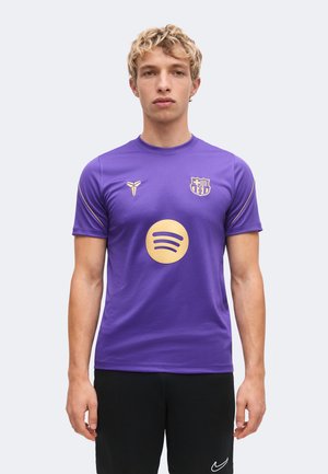 Jeune homme portant un maillot de sport violet avec le logo doré Spotify et le blason du FC Barcelone, debout devant un fond clair uni.