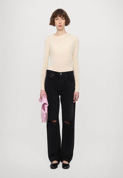 Long-sleeve, light yellow fitted top with textured detailing, paired with distressed black denim jeans and black slip-on shoes, holding pink fabric.