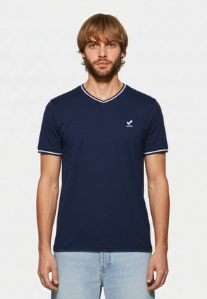 Mann mit hellbraunem Haar und Bart, der ein marineblaues V-Ausschnitt-T-Shirt mit weißem Rand und hellblaue Jeans trägt, steht vor weißem Hintergrund.