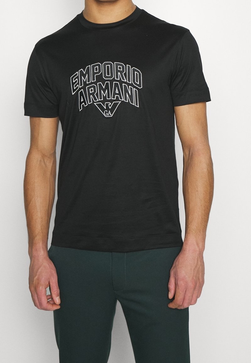 Camiseta de algodón negra con un gran logotipo bordado en blanco de "Emporio Armani" en el pecho. Cuello redondo clásico y mangas cortas.
